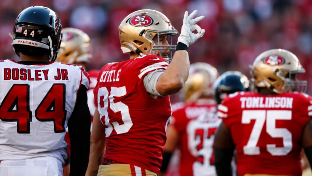 San Francisco 49ers tight end George Kittle celebrates on the field during an NFL game while making his signature hand gesture, with Atlanta Falcons players and teammates in the background.