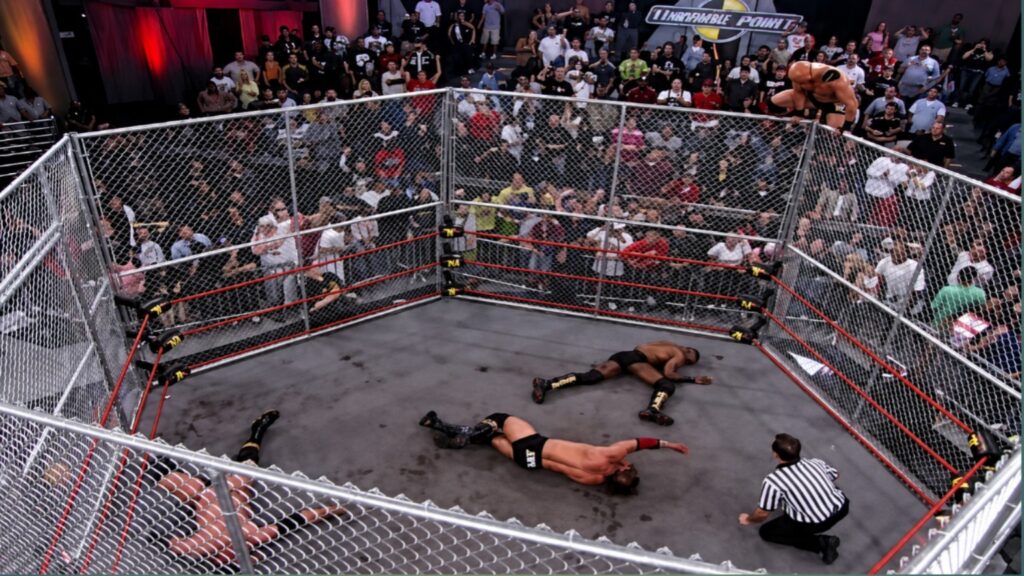 High-angle view of a TNA Lethal Lockdown steel cage match with a wrestler standing on top of the cage as competitors lie inside the ring.