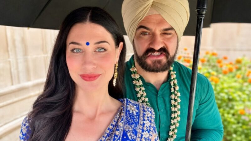 Drew McIntyre and his wife Kaitlyn smiling together in traditional Indian attire, with Drew wearing a green kurta and turban and Kaitlyn in a blue embroidered saree.